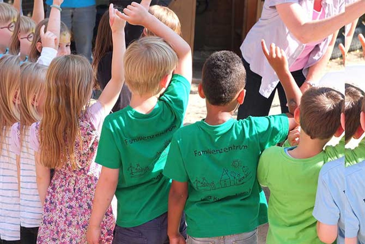 Eine Gruppe Vorschulkinder stehen mit dem Rücken zum Fotografen in einem Halbkreis.  Sie zeigen mit dem Finger auf.  Vor ihnen steht ein Erwachsener, der ihnen eine Frage stellt.