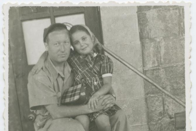 Ein junges Mädchen sitzt auf dem Schoß eines Mannes, sehr wahrscheinlich ihr Vater. Er umarmt das Mädchen, sie lehnt Ihren Kopf an seinen. Beide sehe zufrieden aus. Sie sitzen auf einer Treppe. Das Foto ist in schwarz-weiß.