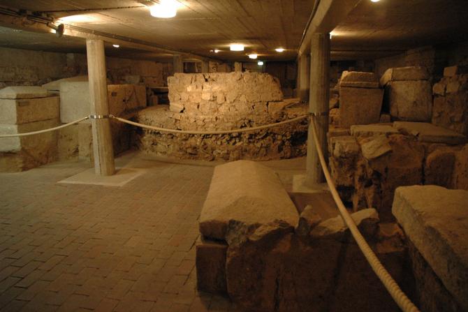 Ein unterirdischer archäologischer Raum mit römischen Mauerresten und Sarkophagen. Der Raum ist schwach beleuchtet, die Überreste sind durch Seile abgesperrt. Zu sehen sind massive Steinquader, runde Fundamente und rechteckige Sarkophage, die zwischen den tragenden Betonsäulen eines modernen Deckengewölbes eingebettet sind. Die Szene vermittelt eine museale Ausgrabungssituation, bei der antike Strukturen in situ erhalten und ausgestellt werden.