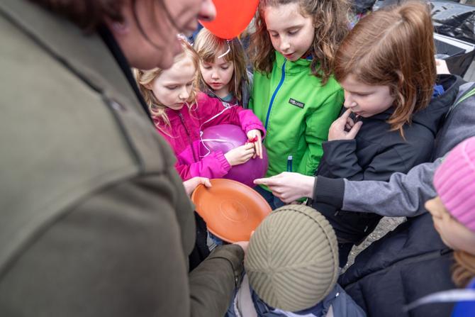 Eine Gruppe von Kindern steht dicht beieinander und betrachtet neugierig einen rotbraunen Teller oder eine Tonschale, die eine erwachsene Person in der Hand hält. Einige Kinder zeigen auf das Objekt, andere halten bunte Luftballons. Die Szene vermittelt eine kindgerechte Führung oder Mitmachaktion, bei der die Kinder archäologische oder historische Gegenstände erkunden dürfen. Der Fokus liegt auf der konzentrierten Interaktion mit dem Objekt.