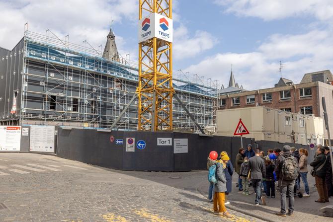 Vor einer großen Baustelle mit Gerüsten und einem gelben Baukran hat sich eine Gruppe von Menschen versammelt. Einige Kinder in der Gruppe halten bunte Luftballons. Im Hintergrund ist ein Gebäude mit dunkler Fassade im Bau, daneben Containerbüros und ältere Backsteinarchitektur. Darüber ragt ein historischer Turm in den teils bewölkten Himmel. Die Szene wirkt wie eine öffentliche Führung oder Veranstaltung im urbanen Raum, bei der sich Interessierte über die Baustelle informieren. 