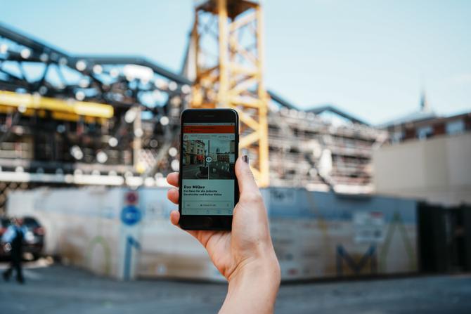 Im Vordergrund hält eine Person ein Smartphone hoch, auf dem eine Website mit dem Titel „Das MiQua – Ein Haus für die jüdische Geschichte und Kultur Kölns“ geöffnet ist. Im Hintergrund ist eine große Baustelle mit Metallgerüsten und einem gelben Baukran zu sehen. Die Kamera fokussiert auf das Smartphone, während der Hintergrund leicht verschwommen ist. Das Bild vermittelt den Eindruck, dass digitale Informationen direkt vor Ort zur historischen Einordnung genutzt werden.