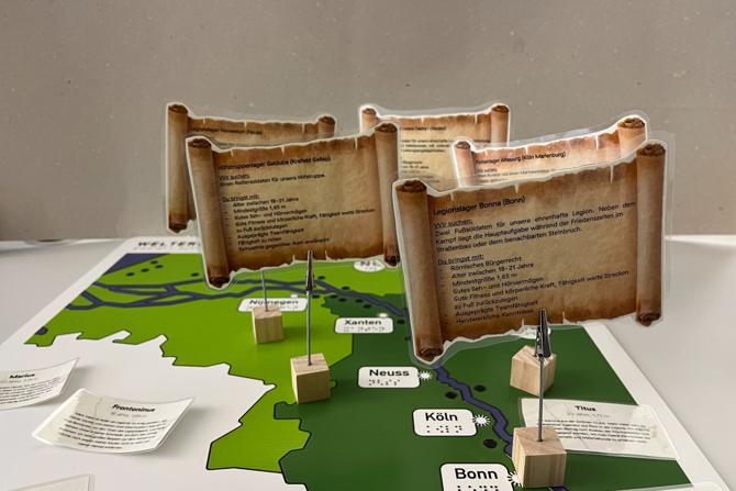 Ein interaktives Lernmodell zeigt eine stilisierte Landkarte mit römischen Standorten am Rhein. Auf der Karte stehen kleine Holzklötze mit aufgesteckten, laminierten Informationsschildern in der Form antiker Schriftrollen. Diese enthalten fiktive „Stellenanzeigen“ für römische Soldaten oder Hilfstruppen mit Anforderungen und Aufgaben. Die dargestellten Orte wie Köln, Bonn, Neuss oder Xanten sind mit Text und Brailleschrift beschriftet. Das Modell lädt spielerisch zur Auseinandersetzung mit dem römischen Militär und dem Leben am Limes ein.