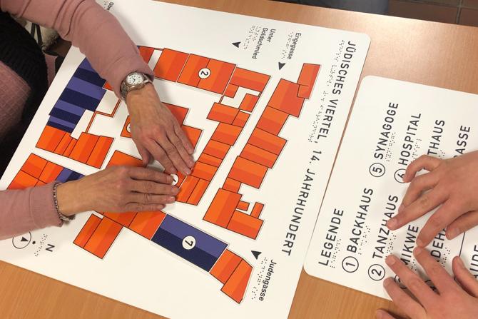 Zwei Personen ertasten ein taktiles Stadtmodell, das auf einem Tisch liegt. Das Modell zeigt einen farbig gestalteten Grundriss eines historischen Viertels mit unterschiedlichen Gebäuden in Orange, Violett und Weiß. Die Personen nutzen ihre Finger, um die erhabenen Strukturen und Beschriftungen zu erkunden. Rechts neben dem Modell befindet sich eine separate Legende mit durchnummerierten Einträgen, die sowohl in Schwarzschrift als auch in Brailleschrift beschriftet ist. Das Modell trägt die Überschrift „Jüdisches Viertel, 14. Jahrhundert“. Die Szene vermittelt eine inklusive Museumssituation, in der sehbehinderte Menschen durch Tasten Zugang zu historischen Informationen erhalten.