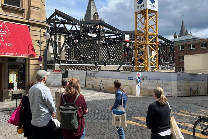 Eine kleine Gruppe von Menschen steht auf einem gepflasterten Platz vor einer großen Baustelle in einer historischen Innenstadt. Eine Person vorne scheint der Gruppe etwas zu erklären. Im Hintergrund ist ein gelber Baukran mit der Aufschrift „Teupe“ zu sehen, dahinter ragen Teile eines alten Fachwerks und Turmspitzen eines historischen Gebäudes in den Himmel. Die Szene wirkt wie eine geführte Tour oder eine Informationsveranstaltung im öffentlichen Raum. Das Wetter ist teils sonnig, teils bewölkt.