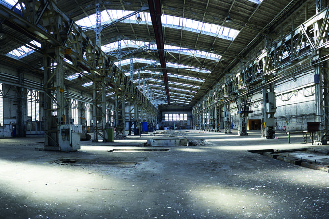 Eine weitläufige, leerstehende Industriehalle mit hoher Decke und Oberlichtern. Die Halle ist von Stahlträgern und Gerüsten durchzogen, Tageslicht fällt durch große Fenster und Dachluken auf den staubigen Boden. Überreste früherer Maschinenanlagen und Gruben deuten auf eine ehemals produktive Nutzung hin. Die Halle wirkt verlassen, aber strukturell intakt, und vermittelt ein Gefühl von industriellem Erbe und vergangener Arbeitswelt.