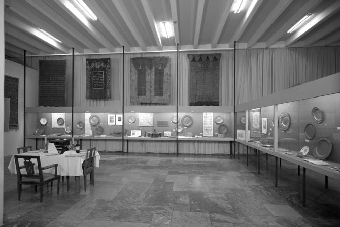 Ein schwarz-weißes Foto zeigt eine museale Ausstellung mit Judaica-Objekten. Entlang der Wände sind in Vitrinen zahlreiche silberne Teller, Ritualgegenstände und gerahmte Dokumente ausgestellt. Darüber hängen reich verzierte, bestickte Textilien mit hebräischen Schriftzeichen. Im Vordergrund steht ein gedeckter Tisch mit vier Stühlen – möglicherweise eine Inszenierung eines festlichen jüdischen Mahls. Die Ausstellung wirkt sachlich und konzentriert, mit einem klaren Fokus auf religiöse und kulturelle Objekte jüdischen Lebens.