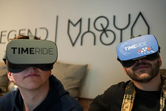 Zwei junge Männer sitzen nebeneinander und tragen Virtual-Reality-Brillen mit der Aufschrift „TIMERIDE“. Eine der Brillen ist zusätzlich mit einem bunten Logo des Projekts „MiQua“ versehen. Im Hintergrund ist eine stilisierte Wandgrafik mit den Schriftzügen „Legend“ und „MiQua“ zu erkennen. Beide Personen wirken konzentriert und sind leicht nach oben gewandt, was auf ein immersives VR-Erlebnis hinweist. Die Szene vermittelt den Einsatz moderner Technik zur Vermittlung historischer Inhalte. 