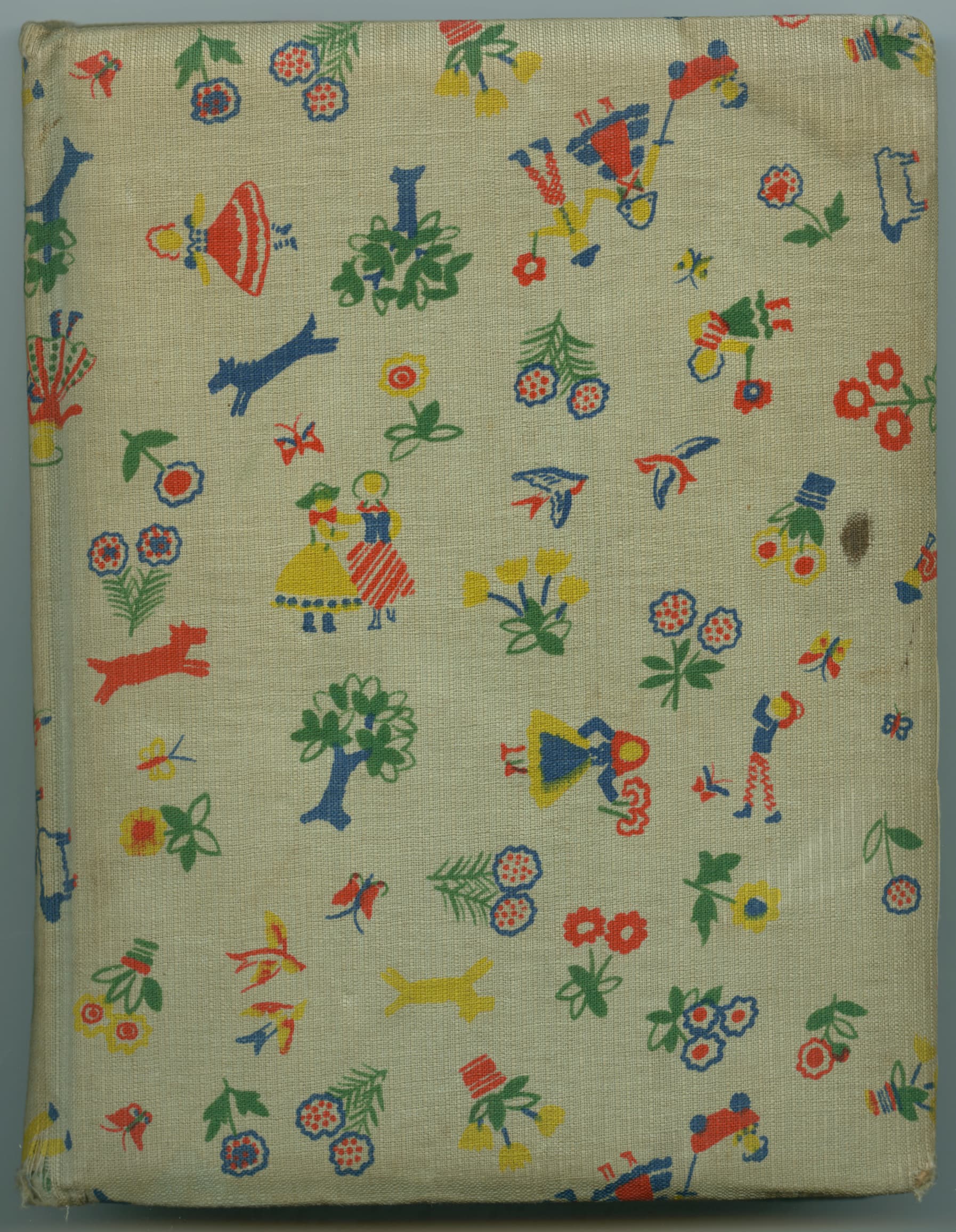 The book cover is made of a piece of fabric with a colorful pattern. The pattern consists of various small, scattered motifs such as birds, flowers, trees and children's toys such as spinning tops and airplanes. The background of the fabric is cream colored and the motifs are in multiple colors such as red, blue, green and yellow.