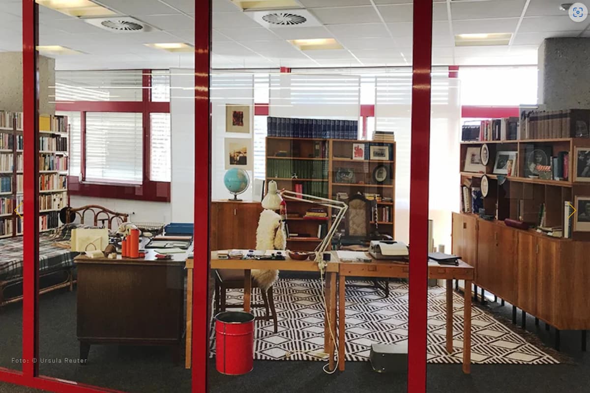 Blick in das Arbeitszimmer mit vielen Büchern im Bücherregal, ein großer schwerer Schreibtisch und ein Globis auf einem Tisch im Hintergrund, Farben rot und weiß und braun
