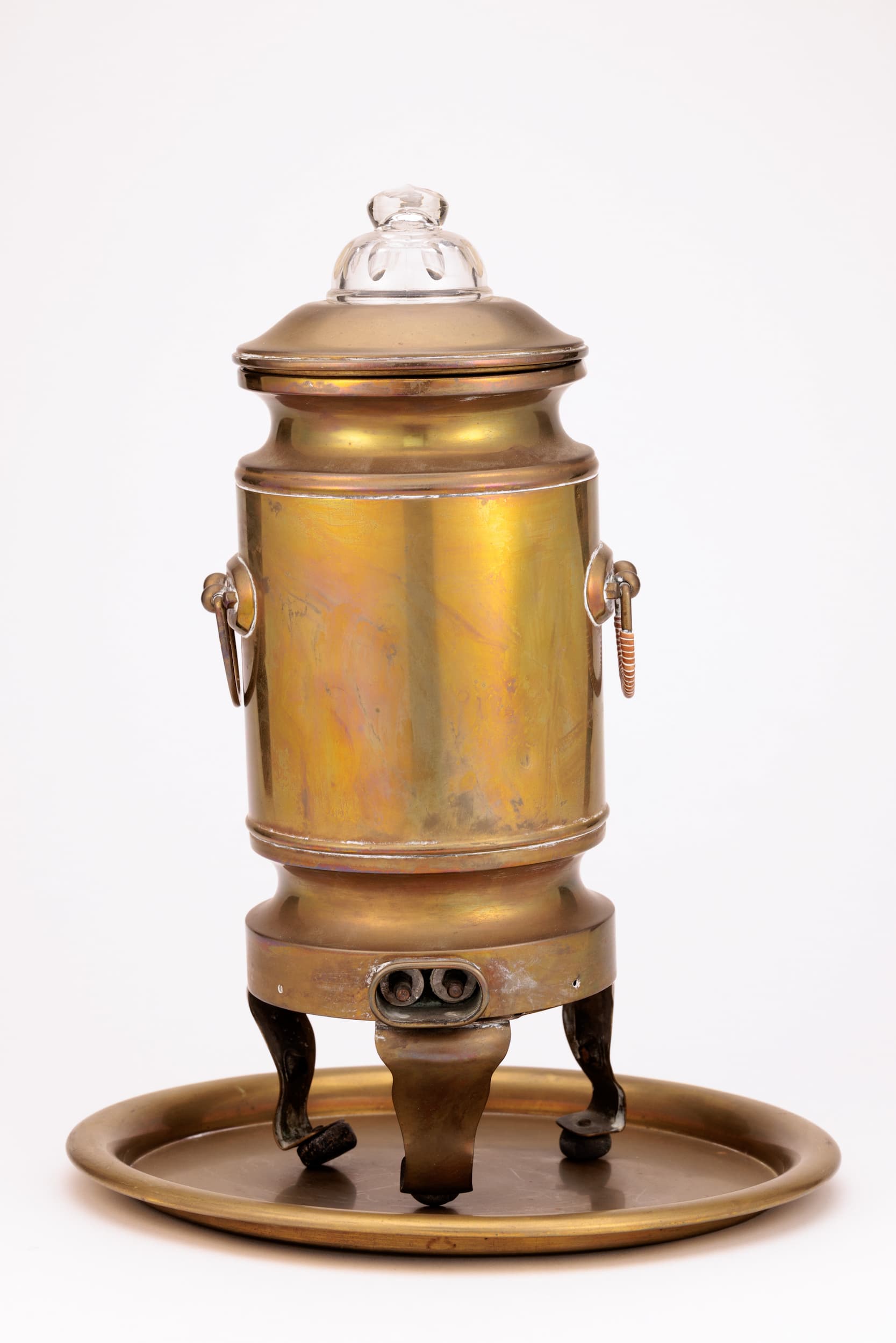 Zu sehen ist ein messing Samovar mit Sicht auf die hintere Seite, unten befindet sich vermutlich ein Stromanschluss, links und rechts mittig sitzt je ein Griff. Der Samovar steht auf 3 Beinen und ist etwas nach hinten geneigt. Er steht auf einem runden Messing Teller. Der Deckel ist mit einem Glasaufsatz geschmückt.