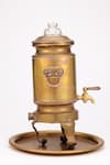Die Seitenansicht eines traditionellen Samowar aus Metall, der häufig zum Erhitzen von Wasser und zur Teezubereitung verwendet wird. Der Samowar ist goldfarben mit kunstvollen Gravuren und steht auf einem passenden runden Tablett. Er hat einen Auslaufhahn nahe dem Boden zum Abgeben von heißem Wasser, zwei seitliche Griffe zum Tragen und steht auf drei geschwungenen Beinen. Der obere Teil hat einen Deckel mit einem Knaufgriff.