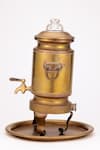 Die Rückansicht eines traditionellen Samowar aus Metall, der häufig zum Erhitzen von Wasser und zur Teezubereitung verwendet wird. Der Samowar ist goldfarben mit kunstvollen Gravuren und steht auf einem passenden runden Tablett. Er hat einen Auslaufhahn nahe dem Boden zum Abgeben von heißem Wasser, zwei seitliche Griffe zum Tragen und steht auf drei geschwungenen Beinen. Der obere Teil hat einen Deckel mit einem Knaufgriff.