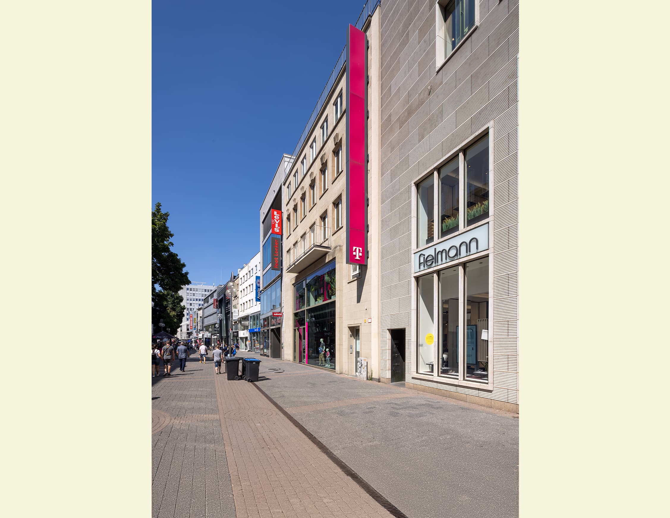 Stadtansicht mit Fußgängern, die auf dem Bürgersteig gehen. Moderne Gebäude säumen die Straße, eines davon trägt das Logo von T-Mobile und ein anderes ein Schild mit der Aufschrift “Telekom”. Der Himmel ist klar und blau, was auf gutes Wetter hinweist.