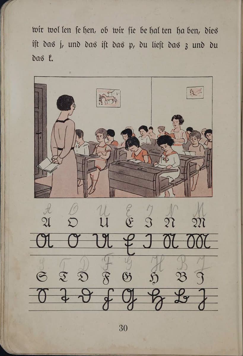 The picture shows a classroom with many children and a teacher standing at the front of the class. One child is reading aloud while the others are following along attentively.  Below are handwritten letters in a grid, and above them are the printed version to teach the children how to write the letters in cursive.