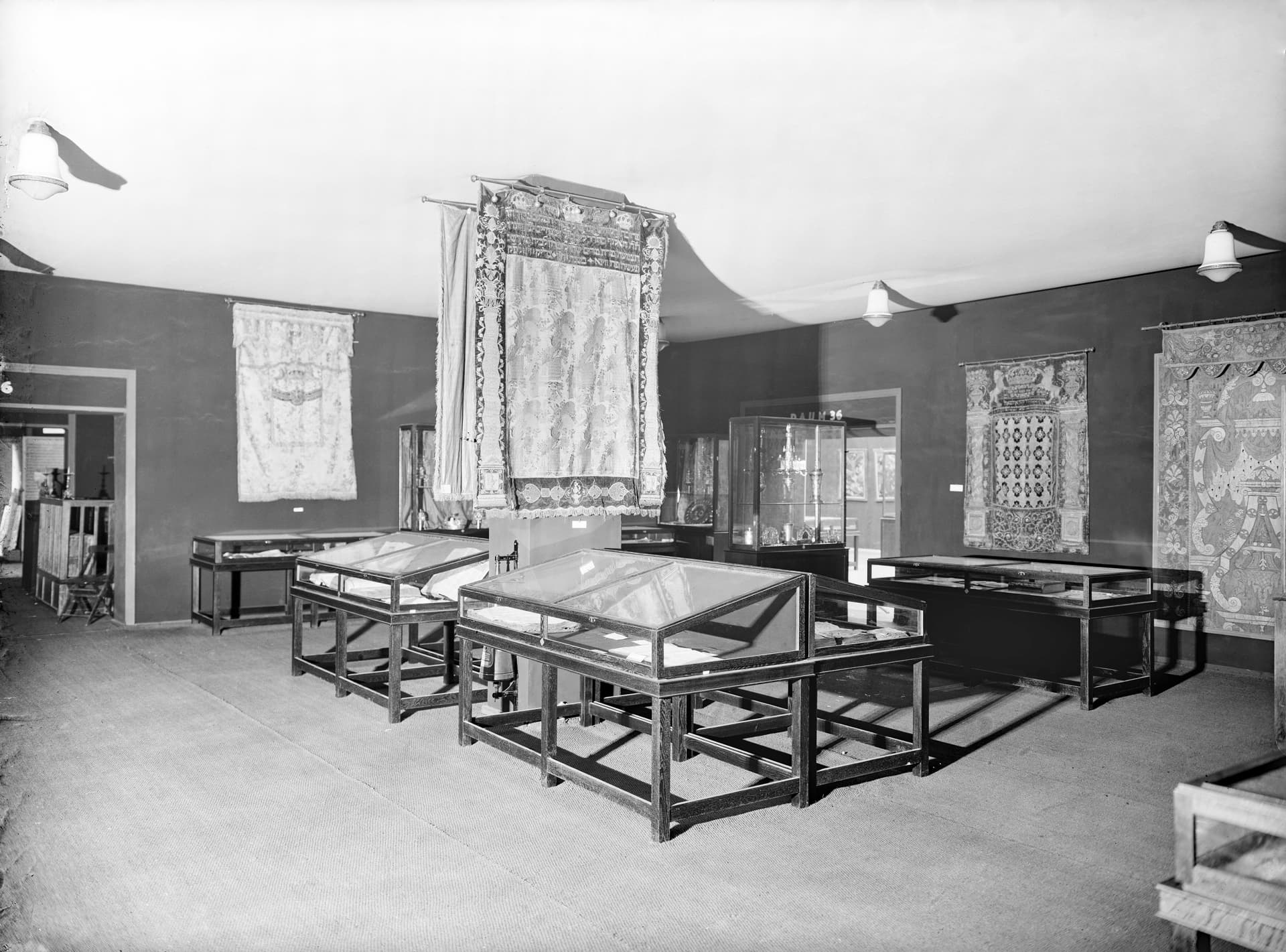 Ein Raum in einem Museum oder einer Ausstellung mit mehreren Glasvitrinen unterschiedlicher Größe. Die Vitrinen enthalten verschiedene Artefakte, die aufgrund der Perspektive nicht genau erkennbar sind. An den Wänden hängen große Textilien oder Wandteppiche, und ein Vorhang ist teilweise vor einem Fenster auf der linken Seite gezogen. Die Decke hat zwei Lichtquellen. Dieses Bild vermittelt Einblicke in die Präsentationsmethoden und den Erhalt von historischen Artefakten.