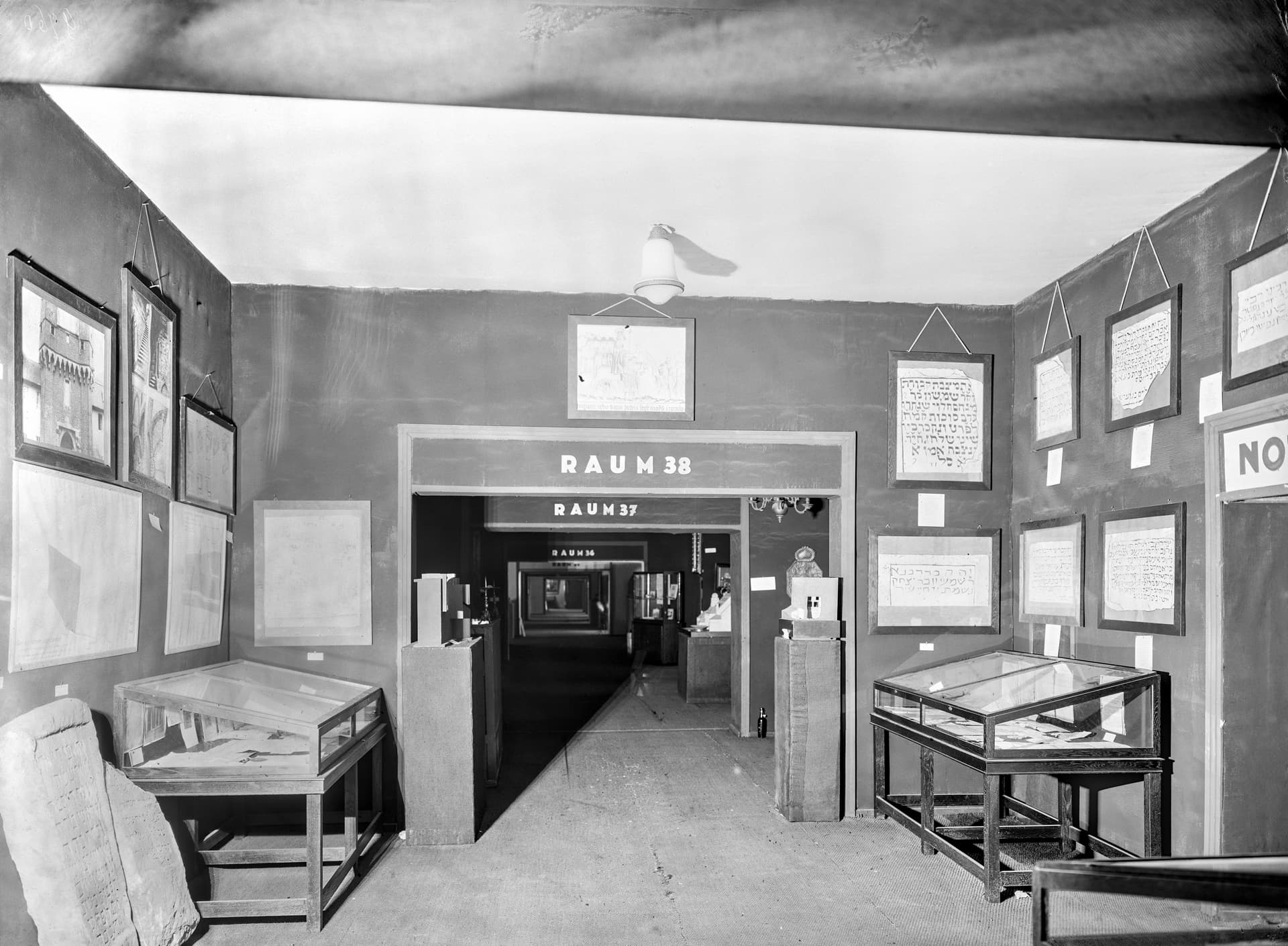 Das Bild zeigt einen Innenraum, möglicherweise einen Raum in einem Museum oder einer Ausstellung. Im Fokus steht eine Tür mit der Aufschrift ‘RAUM 38’, umgeben von verschiedenen gerahmten Texten und Bildern an den Wänden. Im Vordergrund befinden sich Vitrinen, und durch die Tür ist ein weiterer Raum zu sehen. Die Szenerie wirkt historisch aufgrund des monochromen Fotos und des Präsentationsstils.