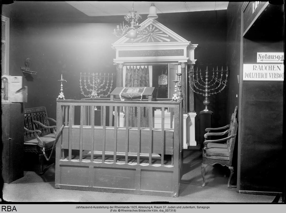 A historic interior of a synagogue with a raised podium, the bimah, surrounded by a wooden balustrade. There are candlesticks on both sides of the bimah. Hanging above the bimah is an elaborate wooden structure, possibly a representation of the tablets of the law. The image is in a monochrome style and depicts a culturally significant scene.