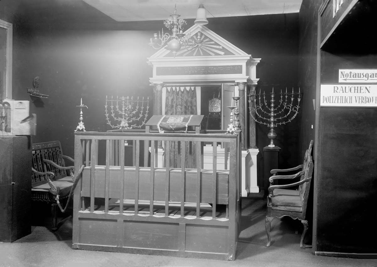 Ein historischer Innenraum einer Synagoge mit einem erhöhten Podium, dem Bimah, umgeben von einer Holzbalustrade. Auf beiden Seiten des Bimahs befinden sich Kerzenleuchter. Über dem Bimah hängt eine kunstvoll gestaltete Holzstruktur, möglicherweise eine Darstellung der Gesetzestafeln. Das Bild ist in monochromem Stil gehalten und zeigt eine kulturell bedeutsame Szene.